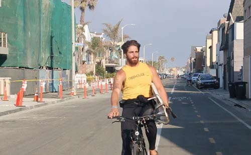 Healthy man rides his bike down Balboa Boulevard with a surf board