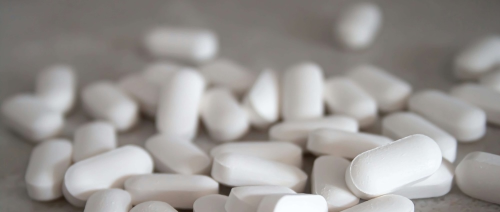 A scattered pile of white oblong Xanax pills lies on a smooth, light-colored surface. The focus is on the foreground, showing detailed textures of the pills, while the background is softly blurred.